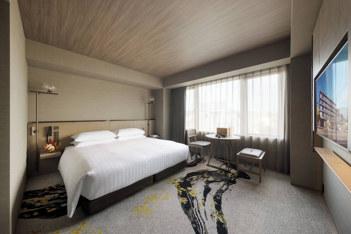Ein Hotelzimmer mit einem großen Bett, Nachttischen, einem Schreibtisch, einem Sessel, einem Fenster mit Vorhängen und Blick auf Gebäude.