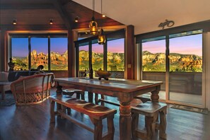 Une pièce spacieuse avec une grande table à manger en bois, de nombreuses chaises et une vue sur un paysage montagneux grâce à de grandes fenêtres.