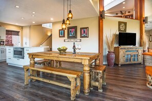 Un coin repas avec une table et des bancs en bois, une cuisine avec des meubles blancs et un téléviseur dans le salon.