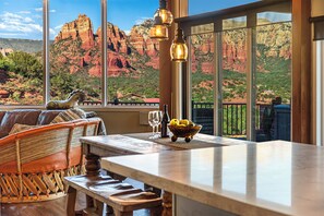 A room with a large window overlooking red rock formations, a wooden table with a bowl of fruit, and a wicker chair.