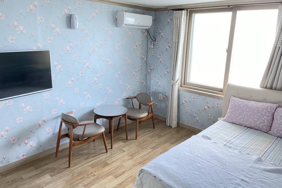 Un dormitorio con cama, dos sillas, una mesita, un equipo de aire acondicionado y una ventana con cortinas.