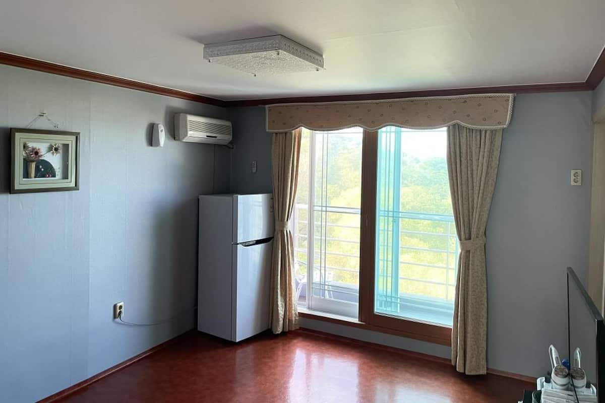 Una habitación con un refrigerador blanco, un respiradero en el techo, una ventana con cortinas y un cuadro en la pared.