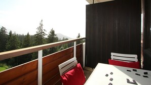 Un balcon doté d’une balustrade en bois, une table blanche avec des coussins rouges, et une vue sur une zone boisée avec des montagnes à l’horizon.