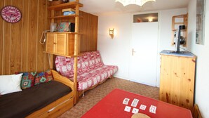 Une pièce chaleureuse avec un lit superposé en bois, un canapé rouge, une petite table avec un jeu de cartes, une armoire en bois et une horloge murale.