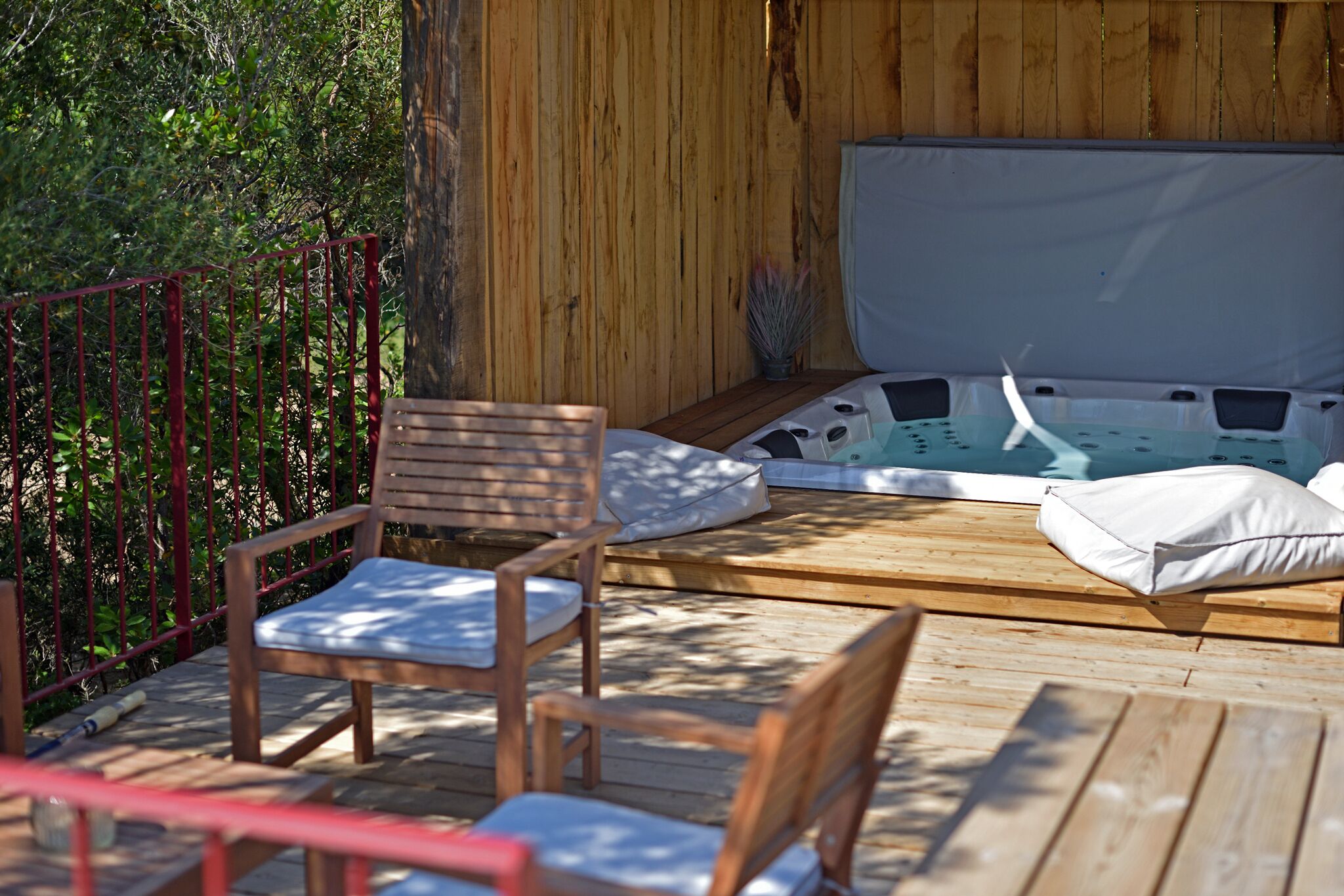 A wooden deck with a hot tub, two wooden chairs, and a red railing.