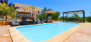 Une piscine entourée d’une terrasse en pierre, agrémentée de chaises longues et de parasols, et une maison avec de grandes fenêtres.