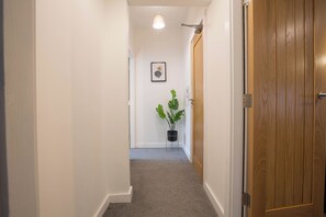 Un pasillo estrecho con una puerta de madera a la derecha, una planta en maceta y un cuadro en la pared.