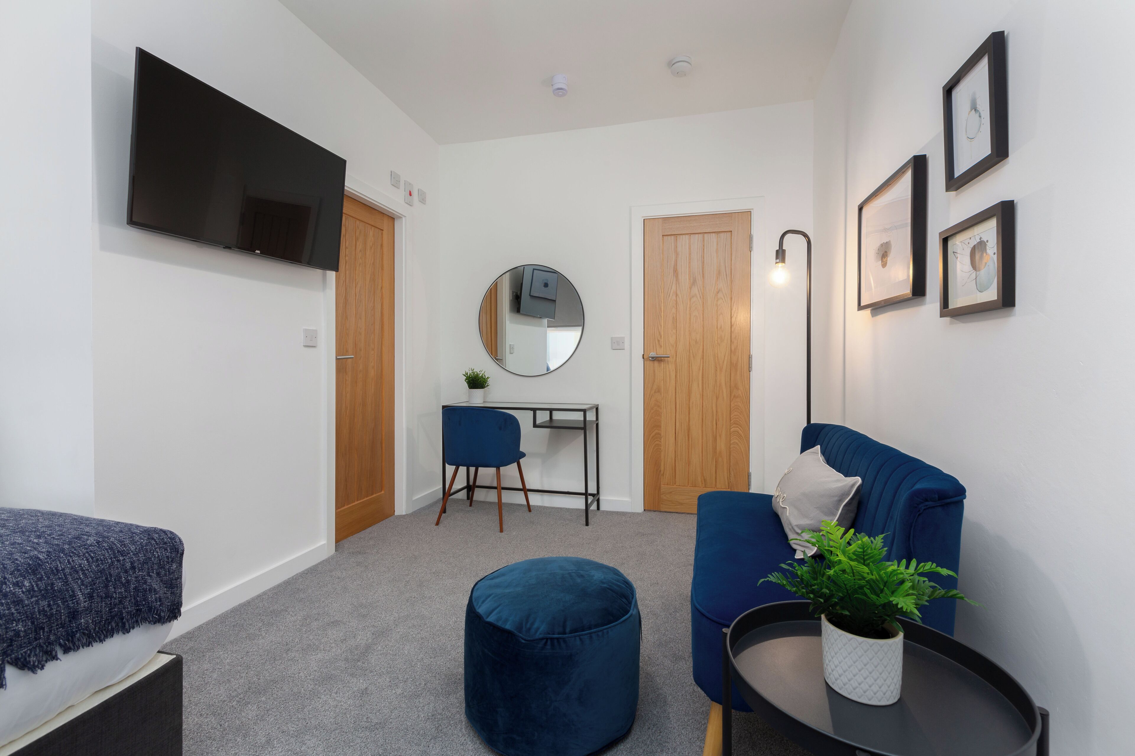 A modern living room with a flat-screen TV, a round mirror, a blue sofa, a small round ottoman, a side table with a plant, and a desk with a chair.
