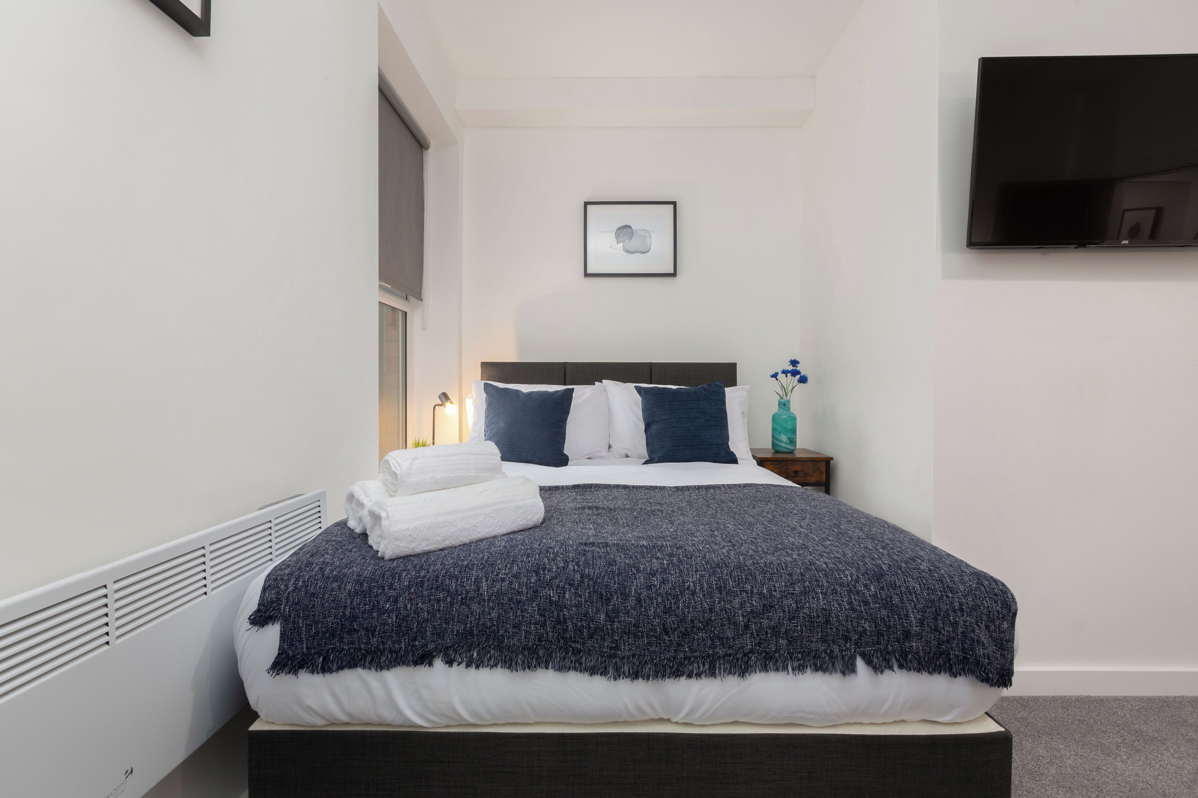 A neatly made bed with a dark blue blanket, white pillows, and a white towel folded on the side. A small bedside table with a teal bottle and a framed picture hangs on the wall.