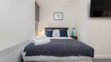 A neatly made bed with a dark blue blanket, white pillows, and a white towel folded on the side. A small bedside table with a teal bottle and a framed picture hangs on the wall.