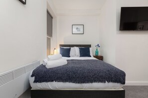Una cama bien tendida con un edredón azul oscuro, almohadas blancas y una toalla blanca doblada al lado. Una mesita de noche pequeña con una botella verde azulada y un cuadro enmarcado cuelga en la pared.