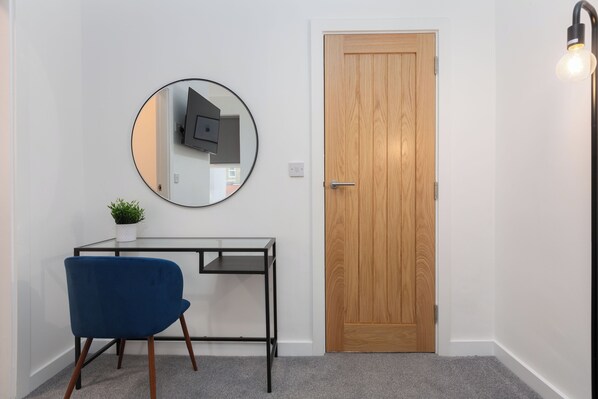 Una habitación con puerta de madera, un espejo redondo en la pared, una mesita con una planta y una silla azul.