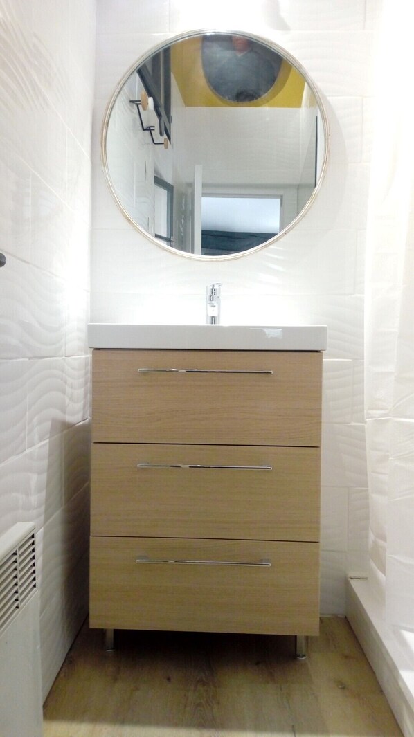 A bathroom with a round mirror, white tiled walls, and a wooden vanity with three drawers.