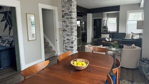 Un salon moderne avec un canapé foncé, une table basse et une table à manger avec des chaises. Un escalier mène à un autre niveau, et un mur en briques apporte une touche d’originalité.