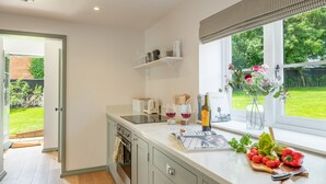 A modern kitchen with a large window, a built-in oven, and a countertop with various items including a cutting board, tomatoes, and a bottle of wine. - Holkham Skies, Wells-next-the-Sea, Norfolk (Wells-next-the-Sea)