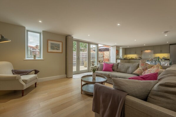 A spacious living room with a large window, a sofa set, a coffee table, and a kitchen area visible through the glass door.