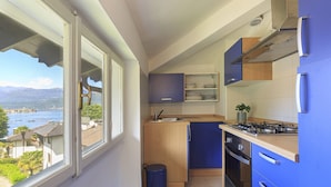 A compact kitchen with a view of a lake and mountains, featuring a sink, stove, cabinets, and a trash bin. - Pandora Stresa (Stresa)
