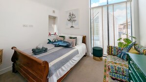 Un dormitorio con una cama, una ventana con persianas, un ventanal pequeño, un enchufe en la pared y un cuadro enmarcado.
