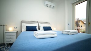 A neatly made bed with blue bedding and white pillows, a bedside table with a lamp, and an air conditioning unit on the wall. - A Queen's room with Balcony (Sorrento)