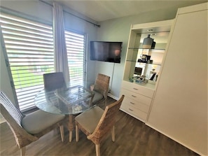 A modern living room with a glass dining table, wicker chairs, a flat-screen TV mounted on the wall, and a built-in cabinet with glass doors.