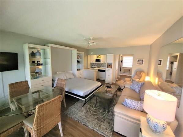 A compact living space with a bed, a sofa, a dining table with chairs, a kitchenette, and a TV mounted on the wall.
