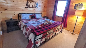Een slaapkamer in een houten blokhut, met een bed, nachtkastjes, een raam met gordijnen en een wandklok.