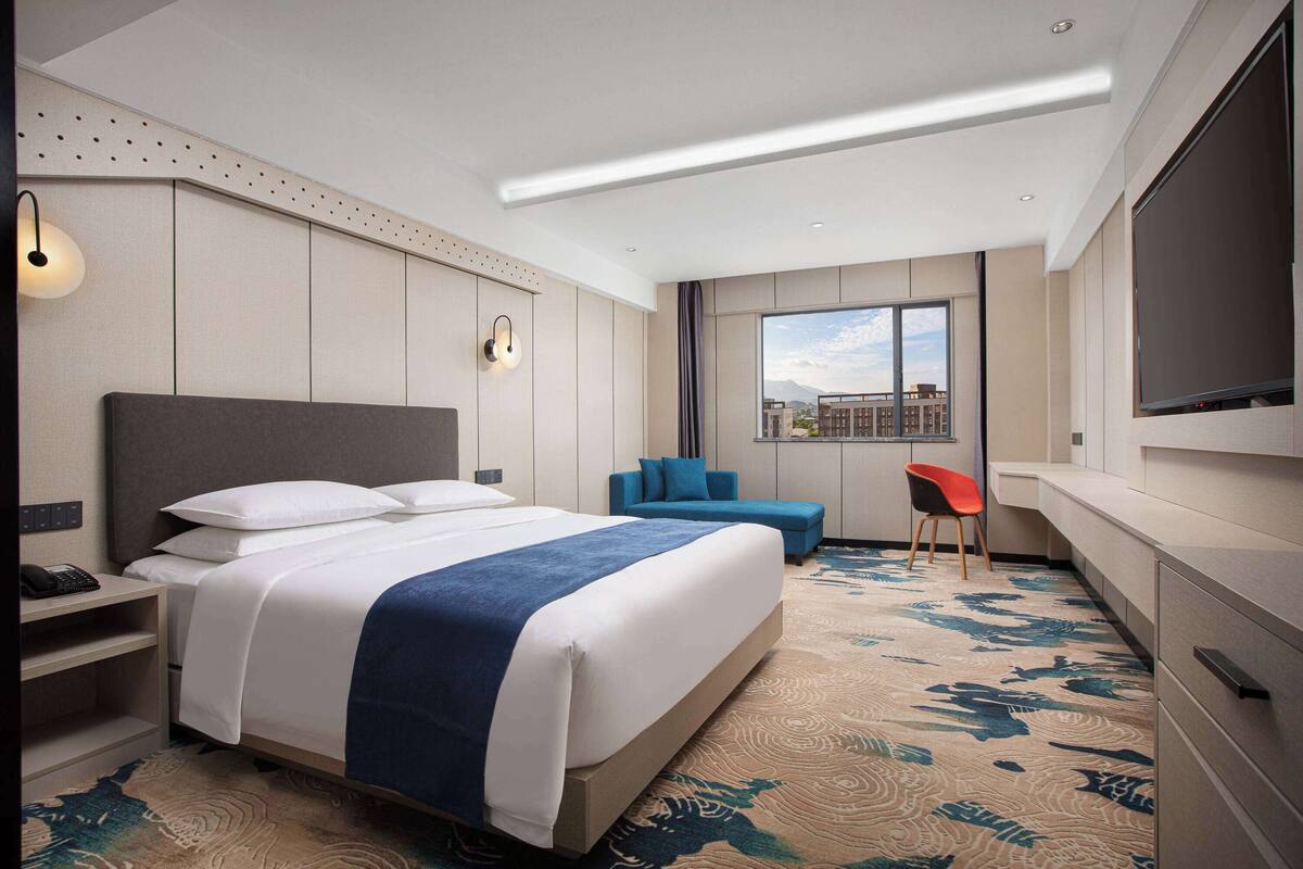 Ein modernes Hotelzimmer mit einem großen Bett, einem Schreibtisch, einem Sessel, einem Fernseher und einem Fenster mit Blick auf die Stadt.