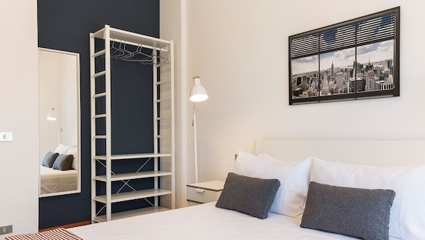 Un dormitorio con cama, armario, mesita de noche, lámpara y una pintura de paisaje urbano.