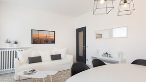 Una sala de estar moderna y bien iluminada, con un sofá blanco, una mesa de centro y un comedor con una mesa redonda. Destacan lámparas colgantes de diseño geométrico, un cuadro de un paisaje urbano y un espejo grande en la pared.