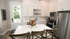 Une cuisine avec des armoires blanches, un réfrigérateur en acier inoxydable, une table à manger avec des chaises et un panier de fruits sur le plan de travail central.