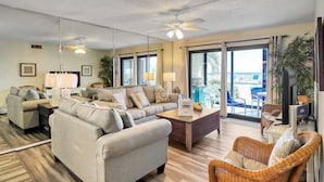 A spacious living room with a sofa set, a coffee table, and a television. There are large windows and a balcony with outdoor seating. - 2 Bedroom Deluxe Gulf Front, Steps to Beach and Pool! Sleeps 7,  First Floor Con (Panama City)