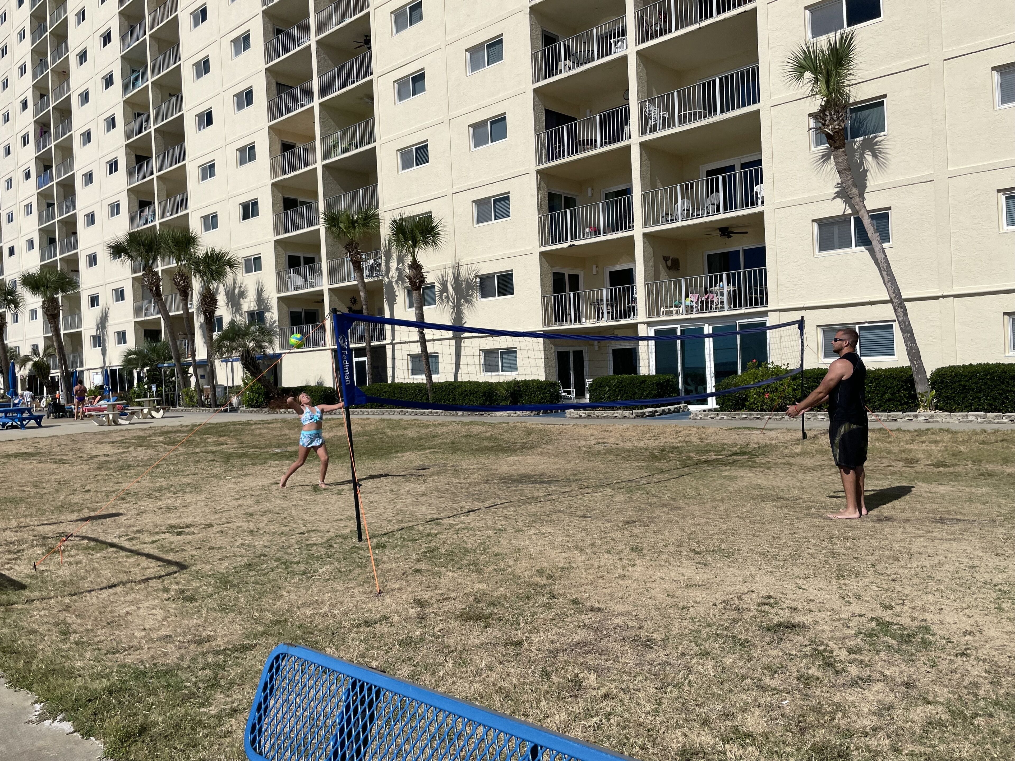 Un immeuble résidentiel à plusieurs étages, doté de balcons, d’un filet de volley-ball et de palmiers.