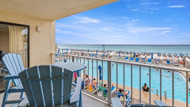 Un balcón con sillas y vista a una piscina frente a una playa con sombrillas y gente.