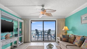 Una habitación con televisión, ventilador de techo, sofá, mesa y vista a la playa a través de la puerta corredera de vidrio.