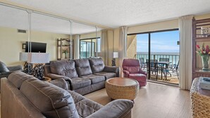 A living room with a sofa, armchair, and coffee table, a TV, and a balcony with a view of the sea.