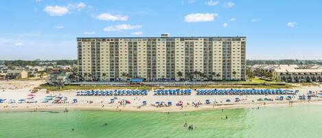 Un grand hôtel à plusieurs étages, une plage en premier plan et de nombreux parasols sur le sable.
