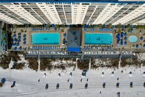 An aerial view of a hotel with two swimming pools, a covered walkway, and a beach area with beach huts.