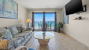 A living room with a sofa, a glass-top coffee table, a flat-screen TV, and a balcony with a view of the sea.
