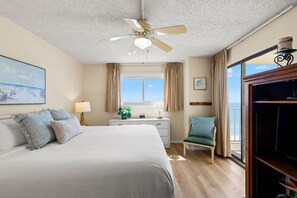 A hotel room with a bed, a desk, a chair, a ceiling fan, a window with curtains, and a view of the ocean.
