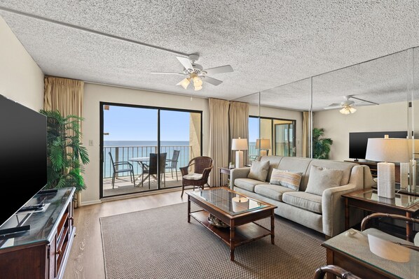 A hotel room with a balcony, a sofa, a coffee table, a TV, and a ceiling fan.