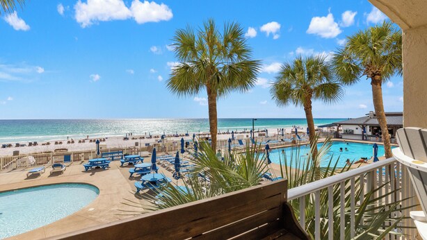 A pool area with lounge chairs and umbrellas, a beach in the background, and palm trees. - 2bd/2ba Condo Ocean views from every room walk in shower FREE BEACH CHAIRS (PCB)