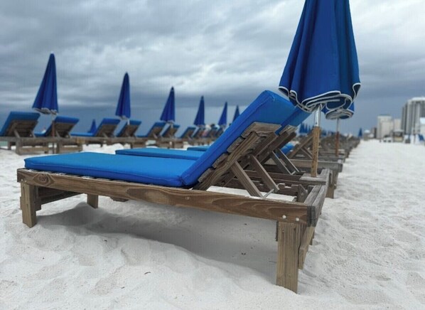 A row of wooden beach chairs with blue cushions and matching blue umbrellas on a sandy beach.