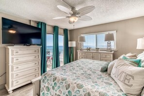Un dormitorio con cama, televisor, cómoda, ventilador de techo, ventana con cortinas y vista al océano.