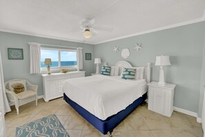 A bedroom with a bed, a chair, a nightstand, a lamp, a window with curtains, and a ceiling fan.