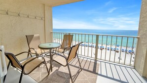 A balcony with a glass table and chairs, overlooking a beach with umbrellas and people.