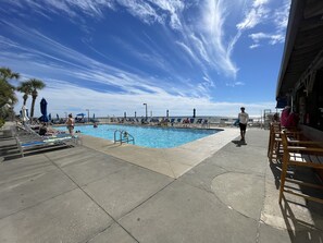 A pool area with lounge chairs and umbrellas, a clear sky, and a view of the ocean.