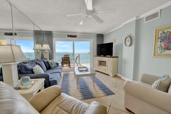 A spacious living room with a sofa, coffee table, and TV, a balcony with a view of the sea, and a ceiling fan.