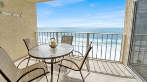 Un balcon avec une table et des chaises en verre, donnant sur une plage sous un ciel dégagé.