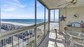 A balcony with a view of the beach and ocean, wicker furniture, and a ceiling fan.