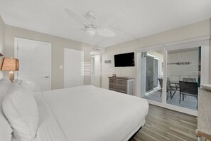 A hotel room with a large bed, a ceiling fan, a TV, a dresser, and a sliding glass door leading to a balcony with outdoor seating.